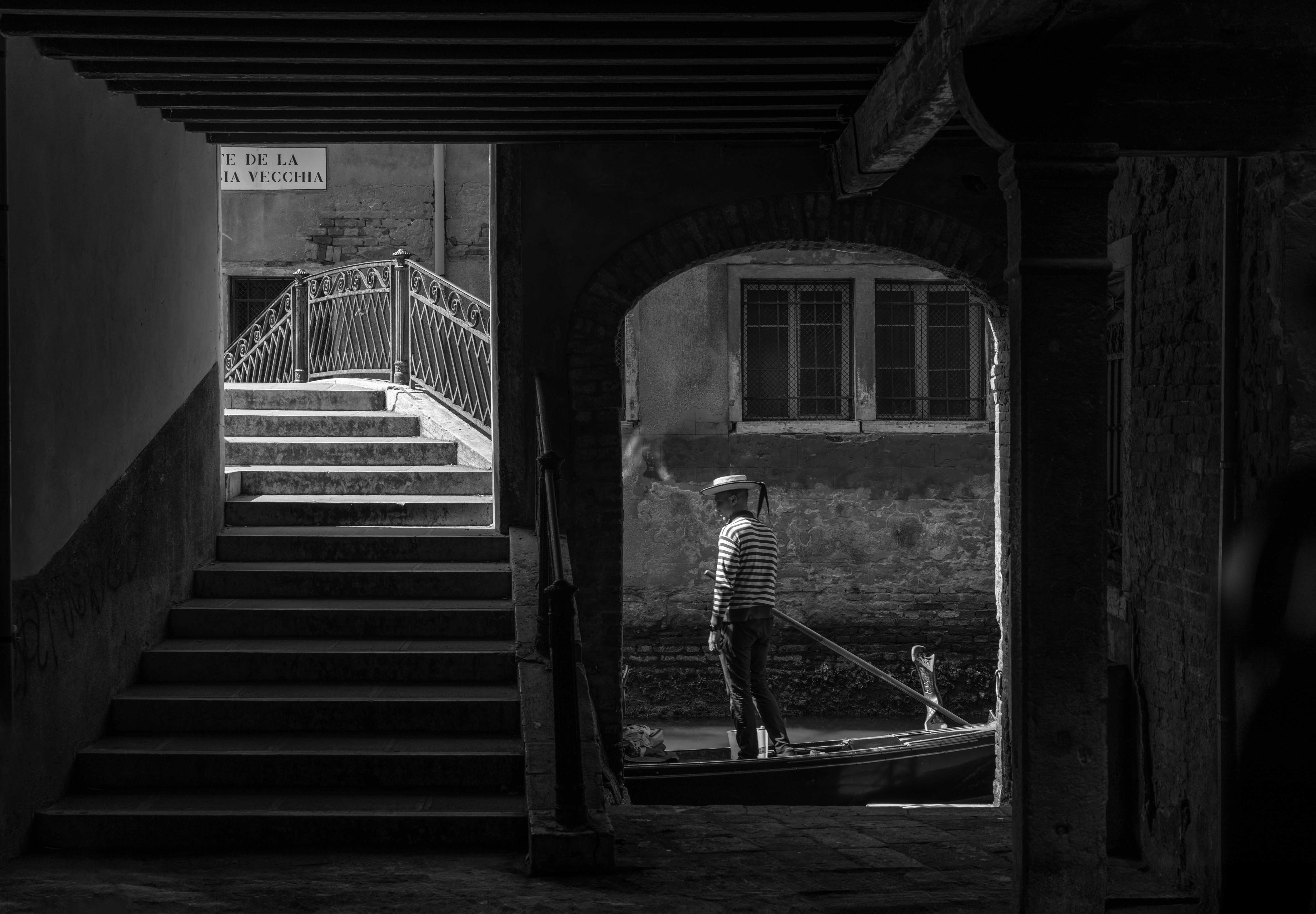 Black-and-white view through a Venetian sotoportego of a striped-shirt gondolier passing beneath an arch beside a stepped bridge